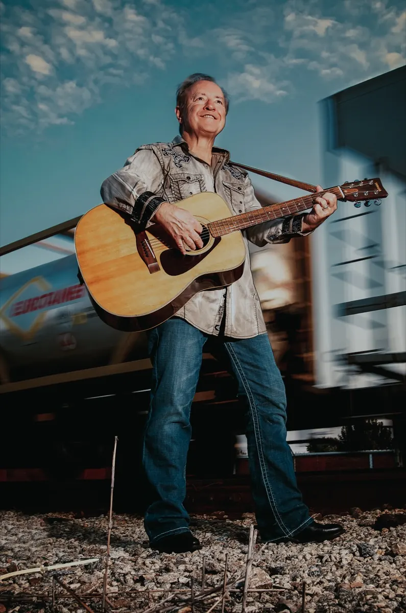 Bobby G. Rice standing with his guitar under a blue sky by a train